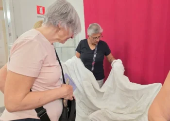 Due donne osservano un ampio tessuto bianco lavorato a sfilato, aperto davanti a un telo rosso, in una sala con vetrine