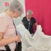 Due donne osservano un ampio tessuto bianco lavorato a sfilato, aperto davanti a un telo rosso, in una sala con vetrine