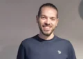 Simone Monello, giovane di Acate, sorride in un ritratto con maglione blu scuro davanti a una parete chiara