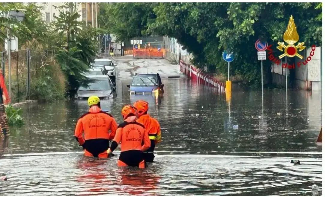 Tre vigili del fuoco in tuta arancione avanzano nell'acqua di un sottopasso allagato, con un'auto blu ferma più avanti