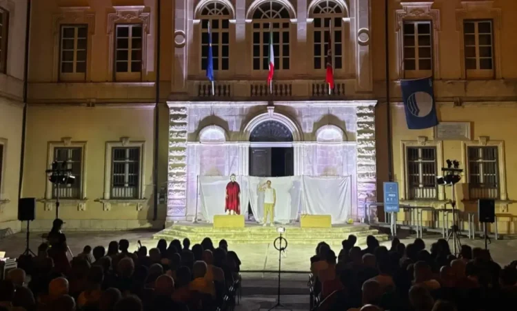 Spettacolo teatrale in piazza Municipio a Pozzallo, due attori sul palco illuminato davanti al pubblico, di sera