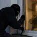 Persona con passamontagna nero tenta di forzare con un piede di porco una finestra illuminata, di notte