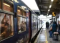 Treno regionale fermo in stazione con passeggeri in attesa sulla banchina coperta, tra valigie e tabellone degli orari