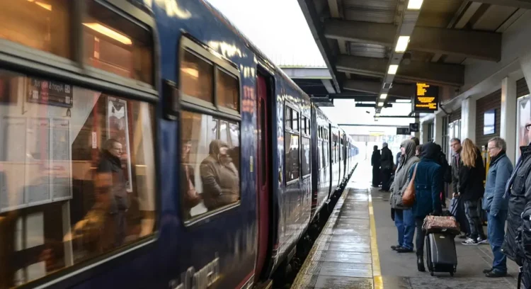 Treno regionale fermo in stazione con passeggeri in attesa sulla banchina coperta, tra valigie e tabellone degli orari