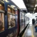Treno regionale fermo in stazione con passeggeri in attesa sulla banchina coperta, tra valigie e tabellone degli orari