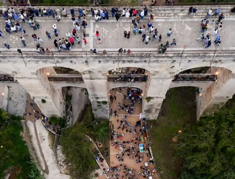 Vista dall'alto del Ponte Vecchio sulla vallata Santa Domenica a Ragusa, con persone sul ponte e nel fondovalle illuminato