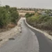 Via 392 a Ragusa, asfalto rattoppato e bordi sterrati tra la vegetazione, sullo sfondo case e traffico