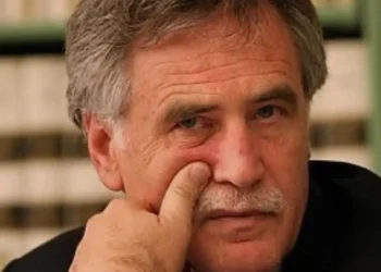 Peppe Drago in primo piano, mano vicino al viso, sfondo di una libreria