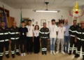 Cinque volontari in gruppo con i vigili del fuoco in divisa, sotto la scritta Vigili del Fuoco, in un ufficio del comando