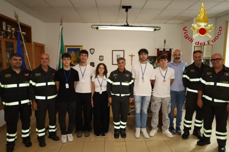 Cinque volontari in gruppo con i vigili del fuoco in divisa, sotto la scritta Vigili del Fuoco, in un ufficio del comando