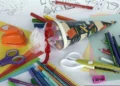 Materiale scolastico colorato: pastelli, pennarelli, forbici, cartoncini e un cono decorato con nastro rosso e tulle bianco