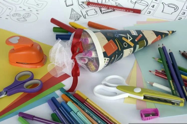 Materiale scolastico colorato: pastelli, pennarelli, forbici, cartoncini e un cono decorato con nastro rosso e tulle bianco