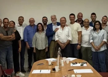 Foto di gruppo in una sala riunioni: uomini e donne in piedi dietro un tavolo ovale con bicchieri, fogli e una bottiglia
