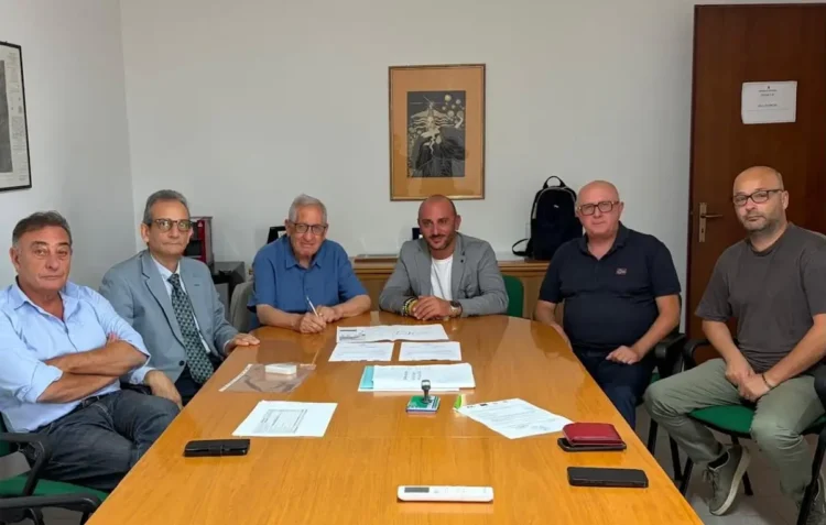 Sei uomini seduti attorno a un tavolo in una sala riunioni, con fogli e un timbro sul piano
