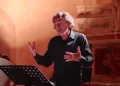 Alessio Boni recita a Ragusa con microfono ad archetto e camicia nera; a sinistra un musicista alla fisarmonica, luce ambrata