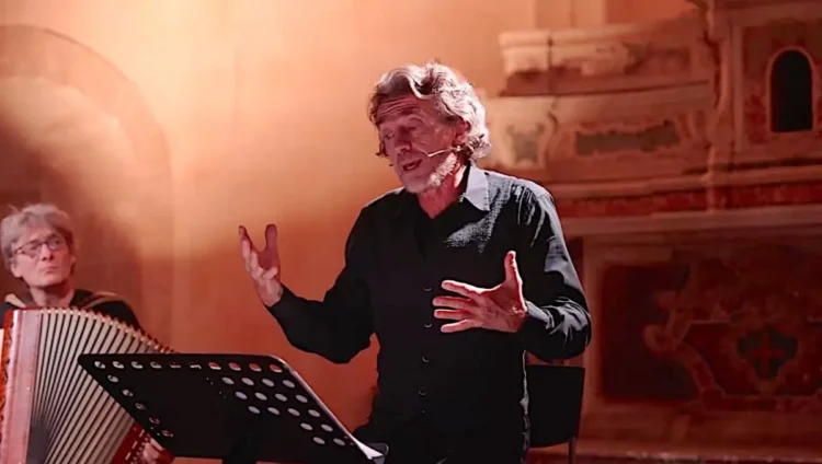 Alessio Boni recita a Ragusa con microfono ad archetto e camicia nera; a sinistra un musicista alla fisarmonica, luce ambrata