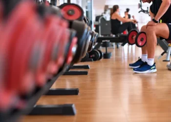 Uomo seduto su una panca in palestra con due manubri in mano, accanto a una rastrelliera di pesi per allenare i muscoli