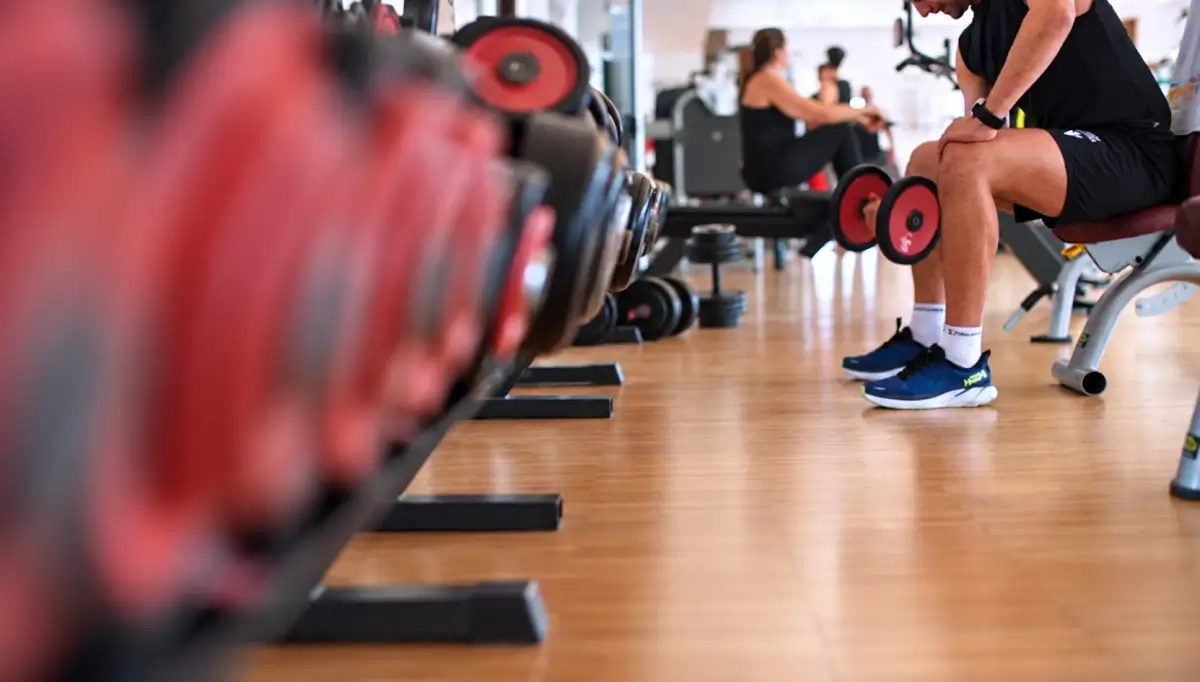 Uomo seduto su una panca in palestra con due manubri in mano, accanto a una rastrelliera di pesi per allenare i muscoli