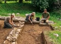 Tre archeologi in ginocchio scavano in una trincea vicino a un muretto di pietre a secco, durante uno scavo archeologico all’aperto