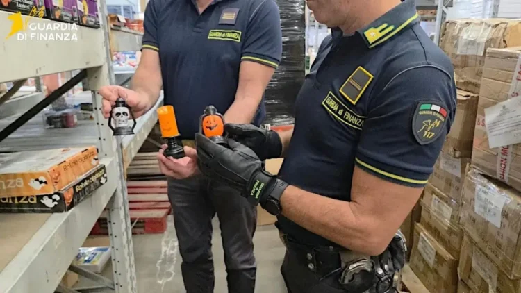 Due finanzieri in un magazzino di Misterbianco mostrano lanterne di Halloween a forma di teschio e zucca tra scaffali e scatoloni