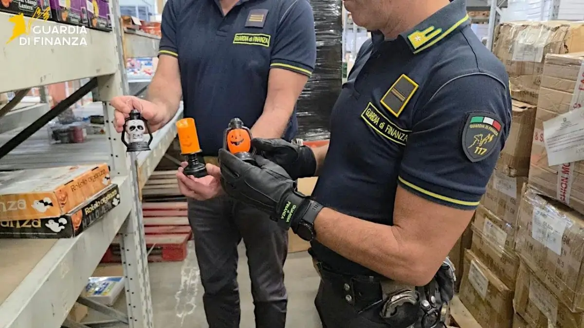 Due finanzieri in un magazzino di Misterbianco mostrano lanterne di Halloween a forma di teschio e zucca tra scaffali e scatoloni