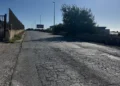 Strada con asfalto crepato e rattoppato a Santa Croce Camerina, tra un muro a secco e una recinzione, con lampioni sullo sfondo