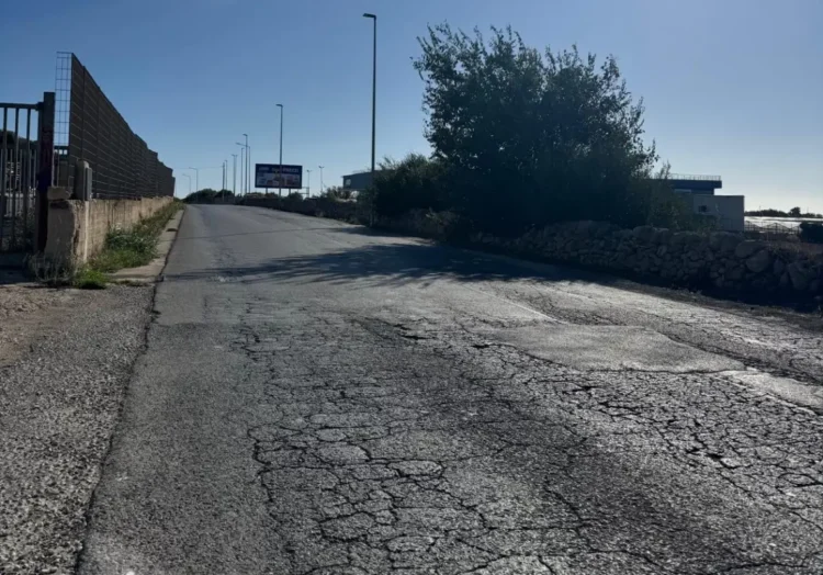 Strada con asfalto crepato e rattoppato a Santa Croce Camerina, tra un muro a secco e una recinzione, con lampioni sullo sfondo