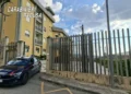 Auto dei Carabinieri con livrea blu e banda rossa parcheggiata davanti a un cancello e a una palazzina gialla