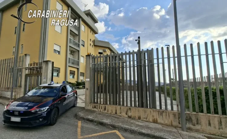 Auto dei Carabinieri con livrea blu e banda rossa parcheggiata davanti a un cancello e a una palazzina gialla
