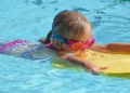Bambina con occhialini rosa nuota in una piscina all’aperto appoggiata a una tavoletta gialla
