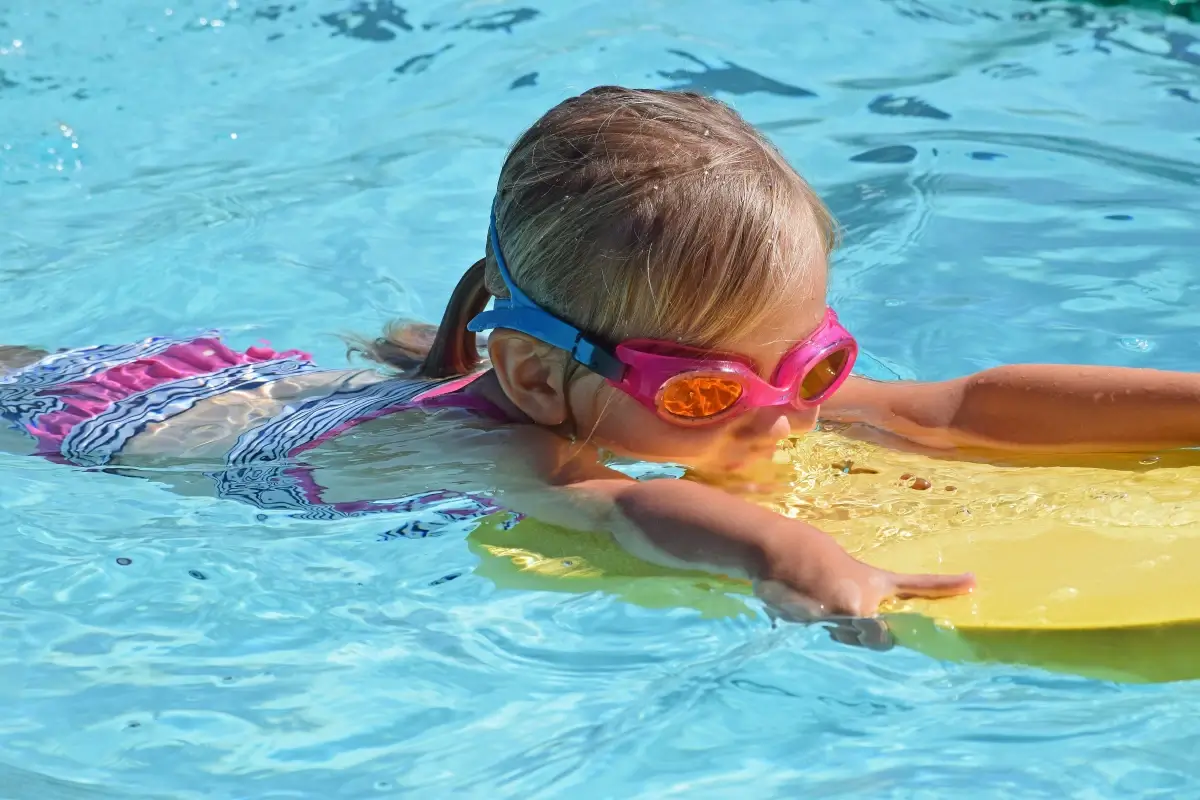 Bambina con occhialini rosa nuota in una piscina all’aperto appoggiata a una tavoletta gialla