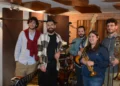 Cinque giovani musicisti in sala prove con tromba, trombone, basso elettrico e bacchette, davanti a una batteria e a un pianoforte