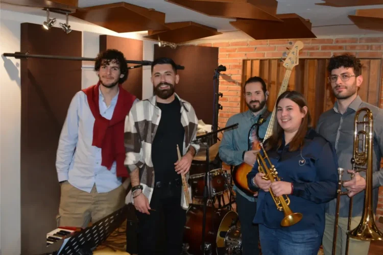Cinque giovani musicisti in sala prove con tromba, trombone, basso elettrico e bacchette, davanti a una batteria e a un pianoforte