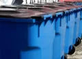 Fila di bidoni carrellati blu con coperchio scuro per l’igiene urbana, allineati davanti a un muro con graffiti