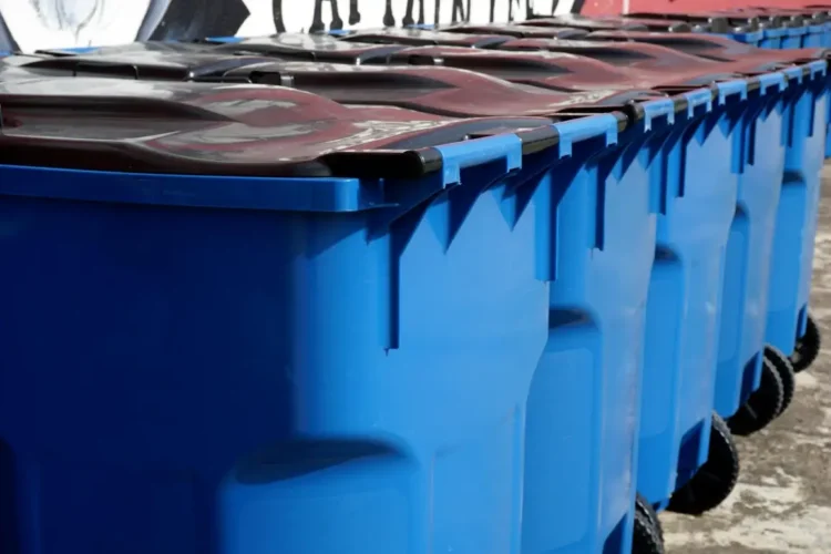 Fila di bidoni carrellati blu con coperchio scuro per l’igiene urbana, allineati davanti a un muro con graffiti
