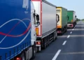 Fila di autotrasportatori con camion e semirimorchi telonati fermi sulla corsia destra di una strada a più corsie