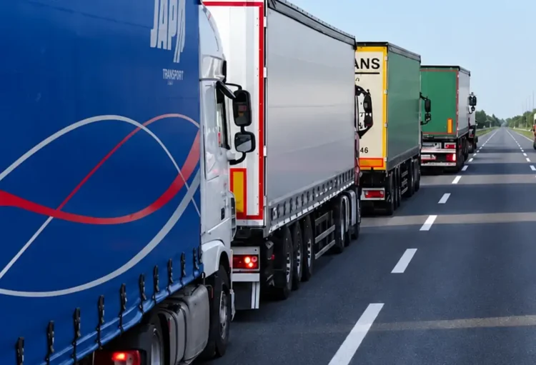 Fila di autotrasportatori con camion e semirimorchi telonati fermi sulla corsia destra di una strada a più corsie
