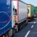 Fila di autotrasportatori con camion e semirimorchi telonati fermi sulla corsia destra di una strada a più corsie