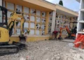 Due miniescavatori sul terreno ricoperto di pietrisco davanti ai loculi del cimitero di Giarratana, con un operaio al lavoro