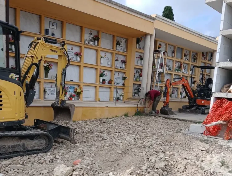 Due miniescavatori sul terreno ricoperto di pietrisco davanti ai loculi del cimitero di Giarratana, con un operaio al lavoro