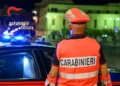 Un carabiniere visto di spalle, con pettorina e berretto arancioni, accanto a un’auto di servizio durante un controllo serale