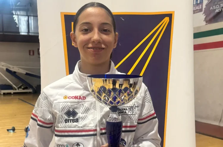 Giovane schermitrice della Conad Scherma Modica in tuta bianca con una coppa argentata, davanti a un pannello blu e giallo
