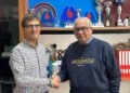 Stretta di mano davanti a uno scaffale con loghi Avis e coppe; uno dei due uomini indossa una felpa Archimede Modica