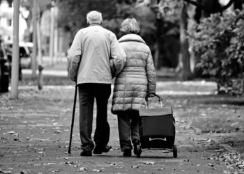 Coppia di pensionati anziani di spalle su un viale con foglie secche, lui con il bastone e lei con il carrello della spesa