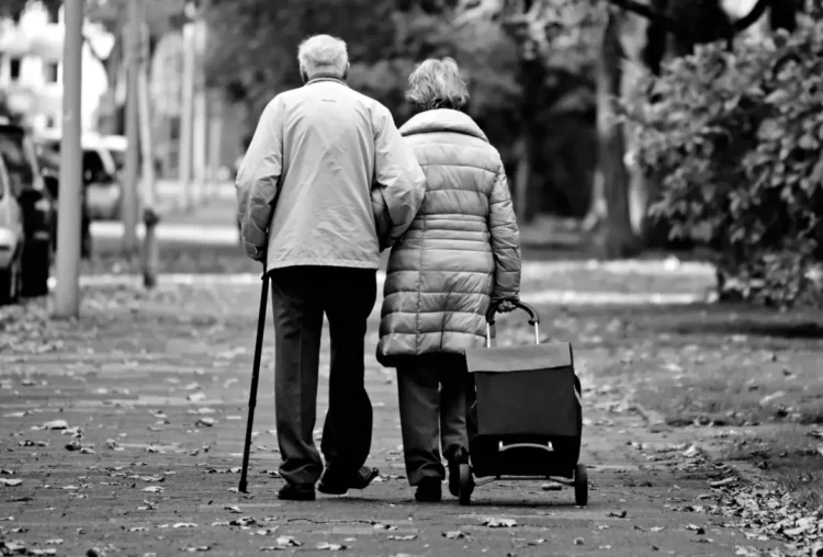 Coppia di pensionati anziani di spalle su un viale con foglie secche, lui con il bastone e lei con il carrello della spesa
