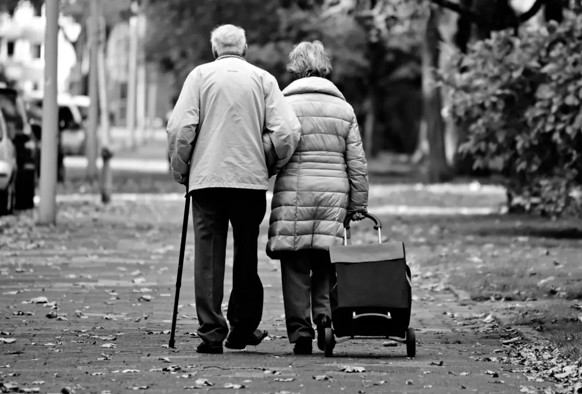 Coppia di pensionati anziani di spalle su un viale con foglie secche, lui con il bastone e lei con il carrello della spesa