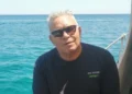 Enzo Ina, capelli bianchi e occhiali da sole, seduto su una barca con il mare azzurro alle spalle