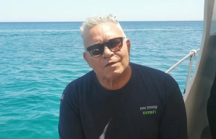Enzo Ina, capelli bianchi e occhiali da sole, seduto su una barca con il mare azzurro alle spalle