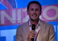 Federico Bennardo, consigliere comunale a Ragusa, parla al microfono in giacca beige davanti a uno striscione blu e rosa