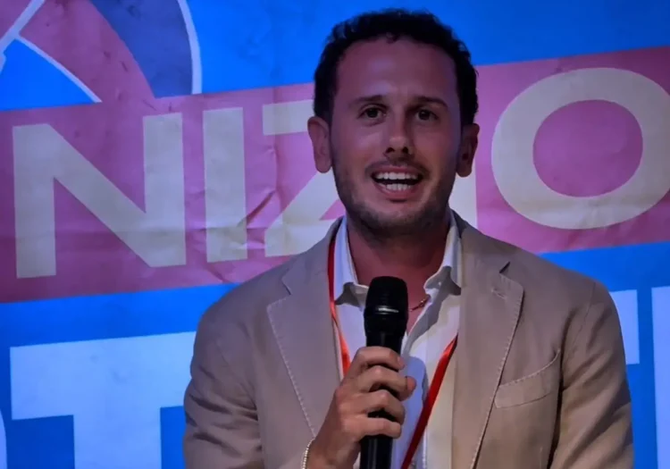 Federico Bennardo, consigliere comunale a Ragusa, parla al microfono in giacca beige davanti a uno striscione blu e rosa